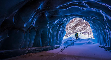A hiker exits a small ice cave while exploring the terminal moraine of Black Rapids Glacier in the winter; Alaska, United States of America.